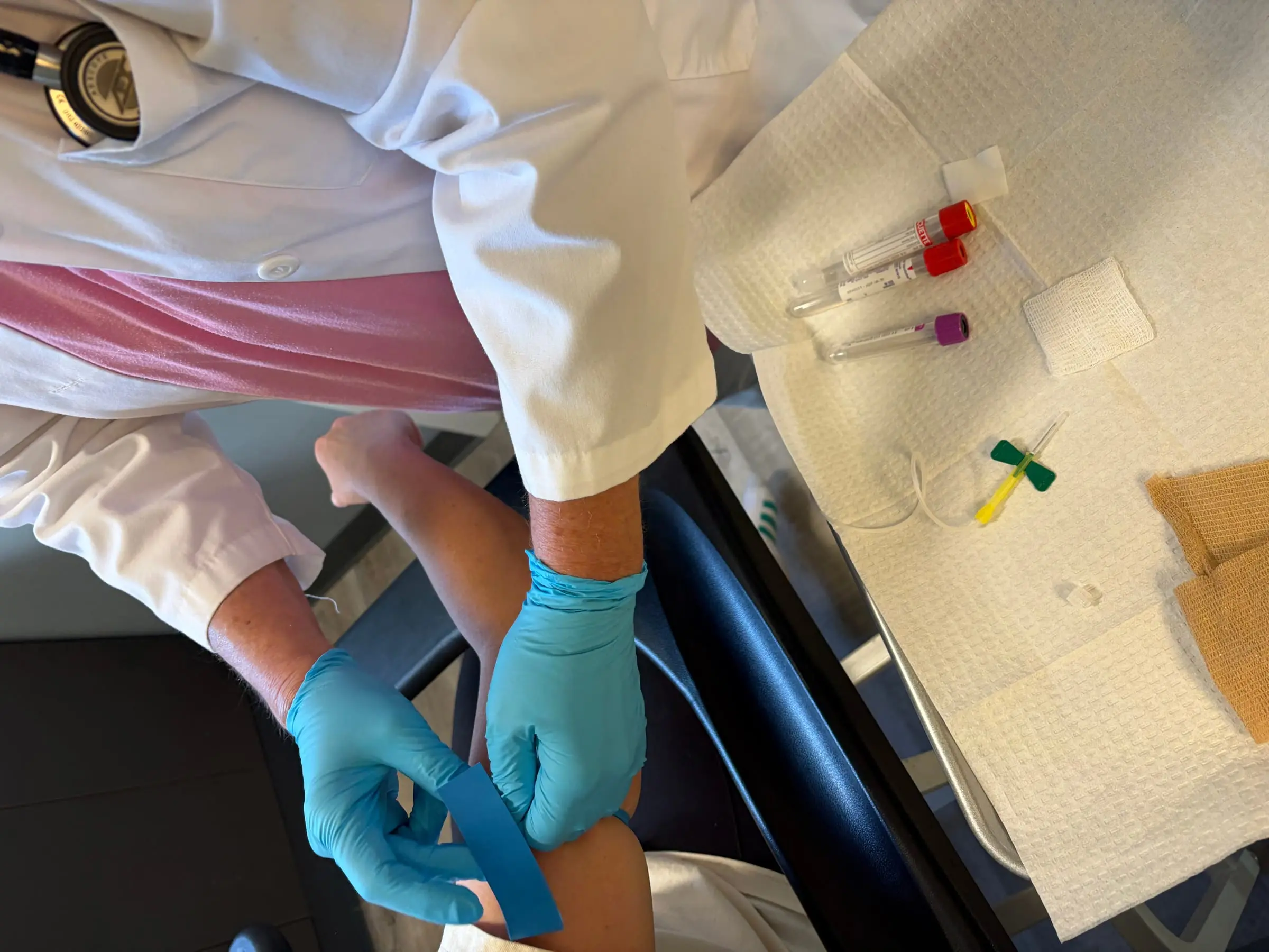 Karen Parmely, NP drawing a comprehensive lab panel — vacutainer tubes, blue gloves, white coat, stethoscope. The first protocol always starts with the labs.