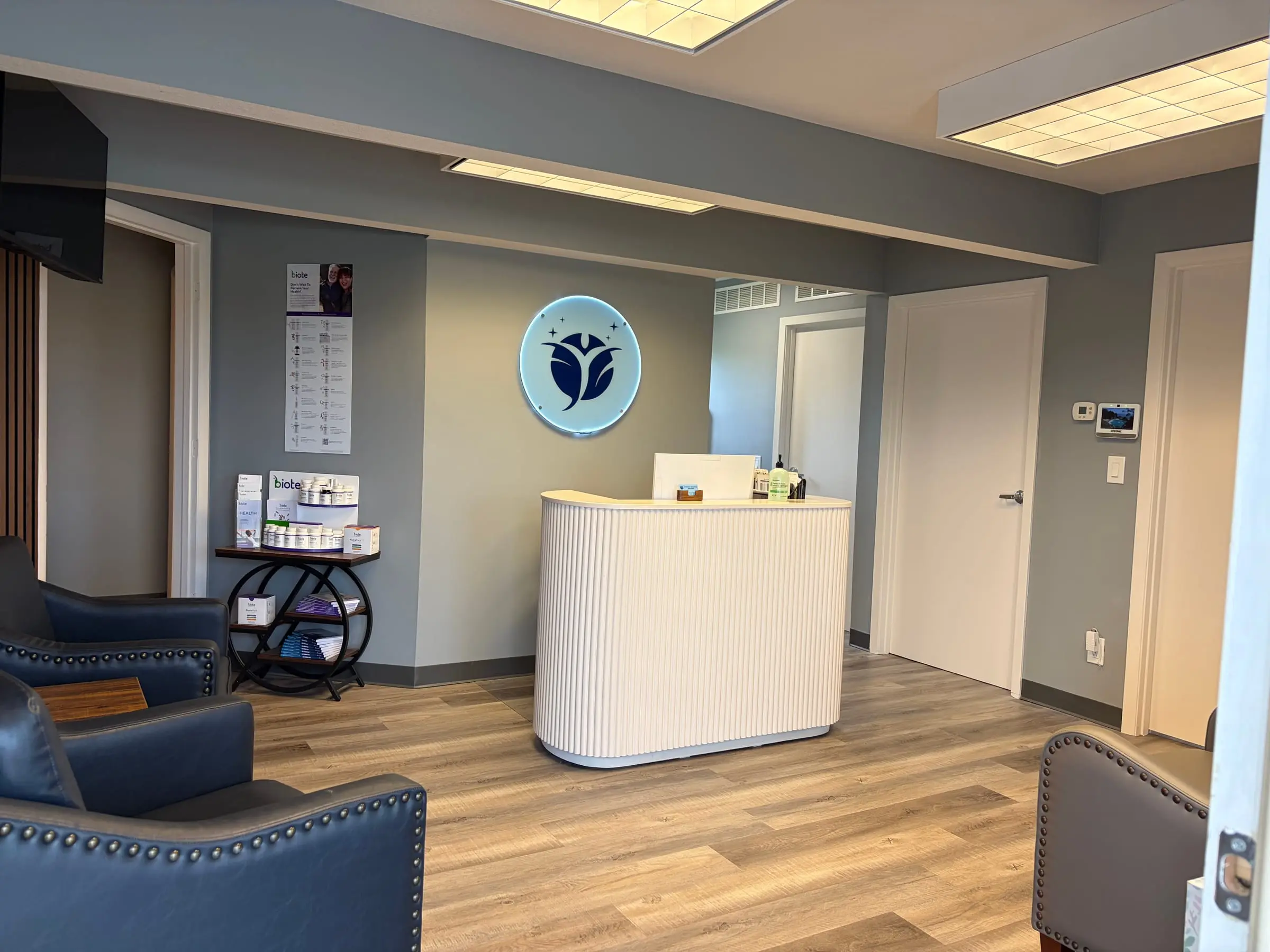 Stilwell Wellness Solutions reception: white fluted desk, backlit lotus logo on the wall, Biote display, navy leather club chairs in the foreground.
